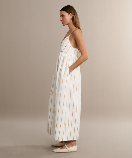 A woman with straight, light brown hair wears the Cove Dress—a long, sleeveless dress with thin gold stripes and delicate shoulder straps—paired with cream flats, against a beige background that highlights the dress’s effortless style.
