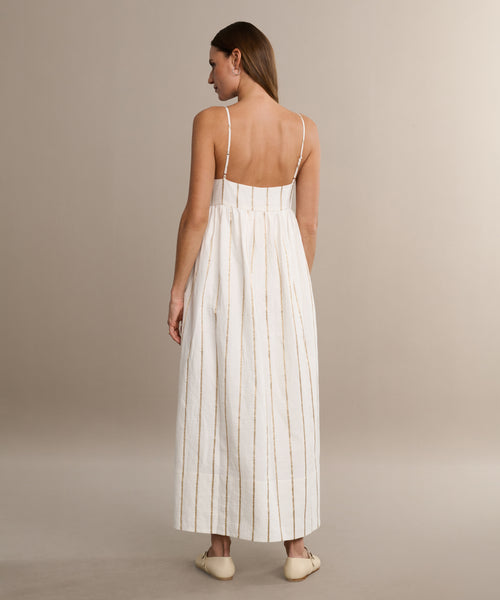 A woman with straight light brown hair wears the Cove Dress, a white casual dress with vertical gold stripes and thin straps, paired with light-colored flats. She faces away against a neutral background.