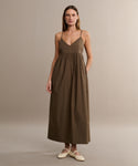 A woman stands against a plain beige background, wearing the Cove Dress—a sleeveless brown cotton design with thin straps and a gathered waist for a relaxed fit—paired with light-colored flat shoes.