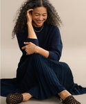 A woman with curly hair sits on the floor, eyes closed and smiling, wearing a navy sweater, pleated navy skirt, gold necklace, and Crochet Jules Mules against a neutral background.