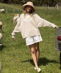 A woman in a cream oversized sweater and white shorts stands on grass near sheep, wearing the Crochet Raffia Sun Hat. She leans casually against a metal object, with trees and a fence in the background.