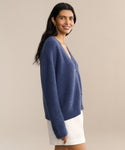A woman with long dark hair, wearing the Cropped Cashmere Cocoon Cardigan in blue and white shorts, stands sideways and smiles against a plain beige background.