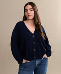 A woman with long brown hair wears the Cropped Cashmere Cocoon Cardigan in navy and blue jeans, standing with her hands in her pockets against a plain beige background.
