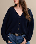 A person wears the Cropped Cashmere Cocoon Cardigan in navy with black buttons, tucked into blue jeans and styled with a black belt featuring a silver buckle. The persons face is partially visible.