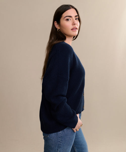 A woman with long brown hair models the Cropped Cashmere Cocoon Cardigan in navy with blue jeans, standing sideways with one hand in her pocket against a plain beige background.