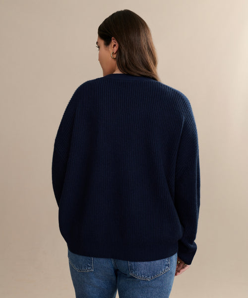 A person with long brown hair, wearing a navy Cropped Cashmere Cocoon Cardigan and blue jeans, stands facing away from the camera against a plain beige background.