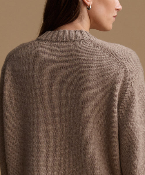 Close-up back view of a woman wearing a ribbed knit sweater in a taupe-brown color, set against a solid warm beige background.