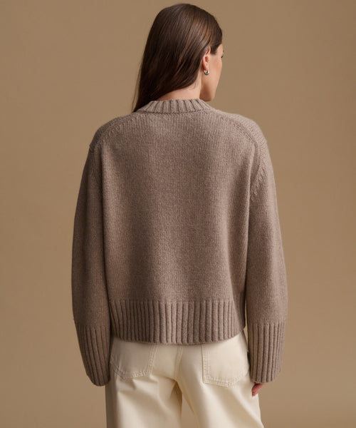 A woman shown from the back wearing a taupe knit sweater and cream trousers against a solid beige studio background.
