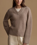 A woman wearing a taupe brown ribbed knit V-neck sweater over a cream top and cream pants, photographed against a solid warm beige studio background.