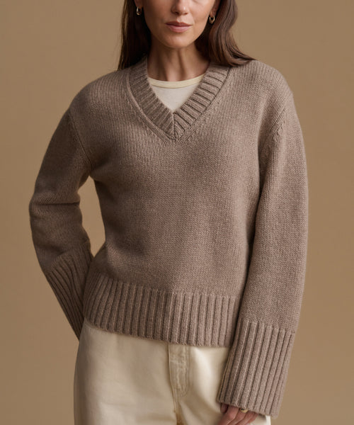 A woman wearing a taupe brown ribbed knit V-neck sweater over a cream top and cream pants, photographed against a solid warm beige studio background.