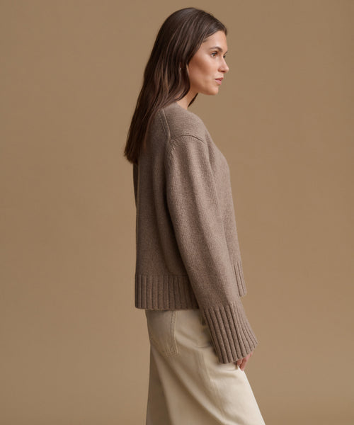 A woman in side profile wearing a taupe knit sweater and cream trousers, set against a solid warm beige studio background.