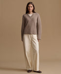 A woman stands against a warm beige studio background wearing a taupe knit V-neck sweater over a cream top, cream wide-leg trousers, and dark brown shoes.