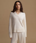 A woman modeling a cream-colored knit sweater with a V-neck and ribbed hem, paired with matching cream pants, against a solid beige studio background.