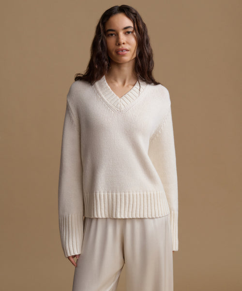 A woman modeling a cream-colored knit sweater with a V-neck and ribbed hem, paired with matching cream pants, against a solid beige studio background.