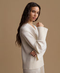A woman modeling a cream-colored ribbed knit sweater with a V-neck and wide sleeves, posed against a solid warm beige studio background.