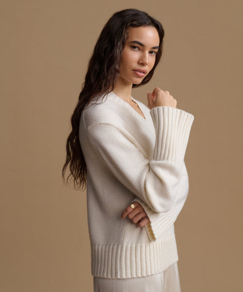 A woman modeling a cream-colored ribbed knit sweater with a V-neck and wide sleeves, posed against a solid warm beige studio background.