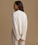 A woman shown from the back wearing a cream-colored knit sweater and matching pants against a solid beige studio background.