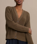 A person wears the Cropped Cotton Cocoon Cardigan—a slouchy, brown ribbed V-neck with large buttons, relaxed fit, and long sleeves—over a dark skirt, standing against a plain beige background.