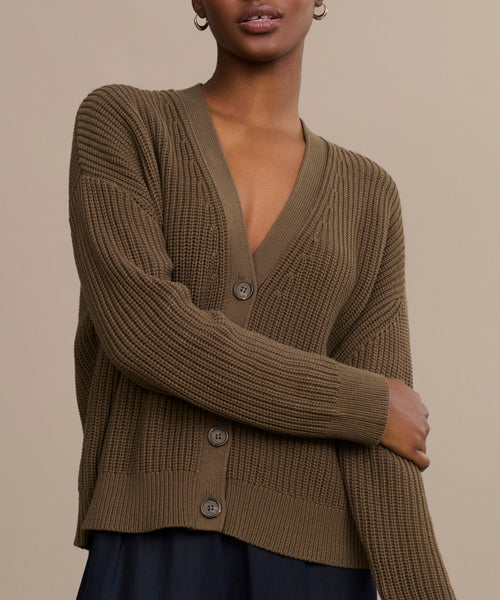 A person wears the Cropped Cotton Cocoon Cardigan—a slouchy, brown ribbed V-neck with large buttons, relaxed fit, and long sleeves—over a dark skirt, standing against a plain beige background.