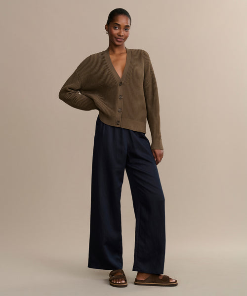 A woman stands against a beige background wearing the Cropped Cotton Cocoon Cardigan in brown, loose navy pants, and brown sandals. One hand is in her pocket, and she looks relaxed.
