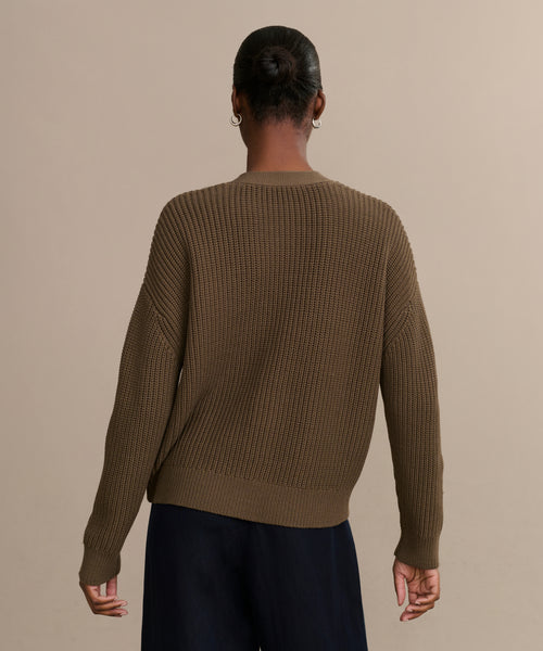 A person with a low bun faces away from the camera, wearing the Cropped Cotton Cocoon Cardigan in olive and dark pants, set against a plain beige background.
