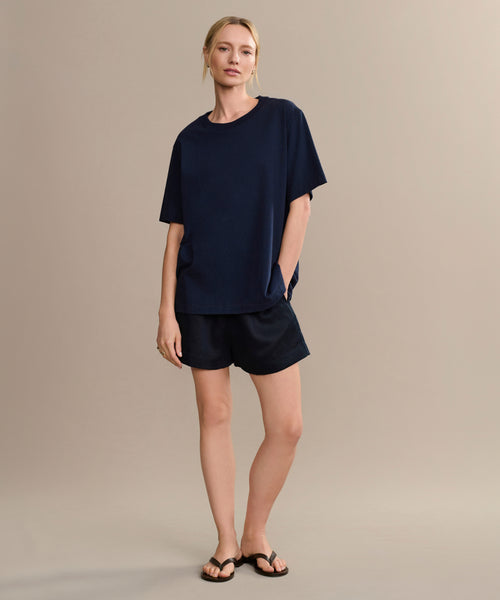 A person with light hair tied back poses casually against a beige background, wearing the Crosby Tee in premium jersey cotton, navy shorts, and black sandals.