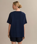 A woman with blonde hair in a low bun is shown from the back, wearing a relaxed fit navy blue Crosby Tee and matching shorts crafted from premium jersey cotton, set against a plain beige background.