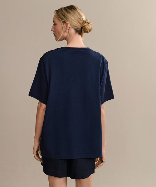A woman with blonde hair in a low bun is shown from the back, wearing a relaxed fit navy blue Crosby Tee and matching shorts crafted from premium jersey cotton, set against a plain beige background.