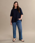 A woman with long brown hair stands against a beige background, wearing the relaxed fit Crosby Tee paired with blue jeans and sandals. One hand is in her pocket while the other rests by her side.