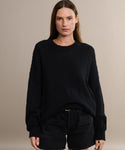 A woman with straight brown hair wears the oversized Cruise Cocoon Crewneck in black and matching black shorts, standing against a plain beige background and looking directly at the camera with a neutral expression.