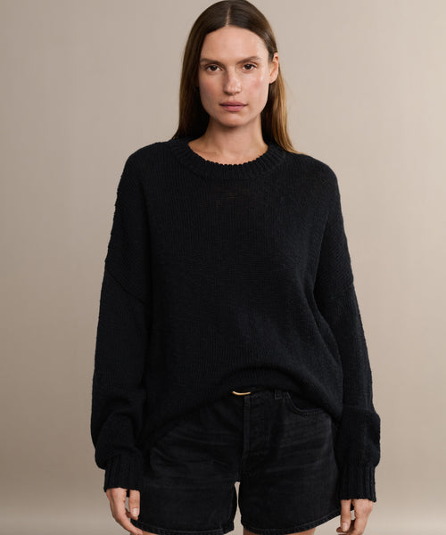 A woman with straight brown hair wears the oversized Cruise Cocoon Crewneck in black and matching black shorts, standing against a plain beige background and looking directly at the camera with a neutral expression.