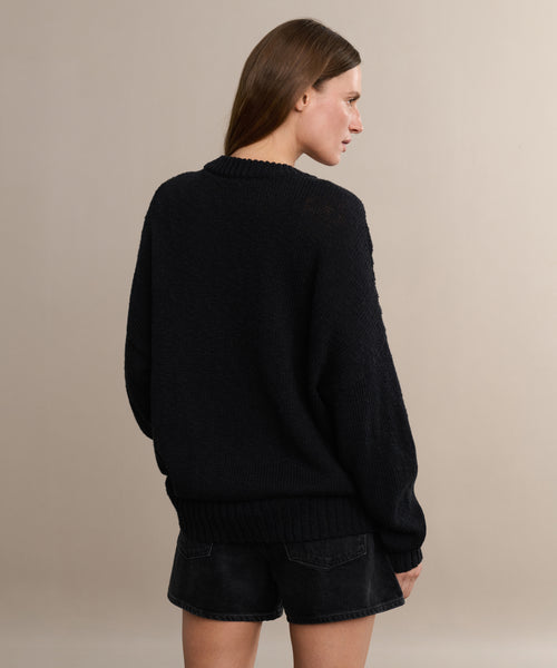 A woman with straight brown hair faces away, wearing the oversized black Cruise Cocoon Crewneck sweater and black shorts against a plain light beige background.