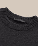 A close-up of the Cruise Cocoon Crewneck, an oversized knit sweater in dark gray with a textured design and ribbed neckline, set against a plain beige background. A black label is sewn inside the collar.