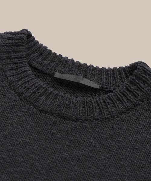 A close-up of the Cruise Cocoon Crewneck, an oversized knit sweater in dark gray with a textured design and ribbed neckline, set against a plain beige background. A black label is sewn inside the collar.