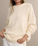 A woman wears the Cruise Cocoon Crewneck, an oversized, cream-colored knit sweater with long sleeves and ribbed crewneck, cuffs, and hem. Her hands are in her white pants pockets, and her face is partially out of frame.