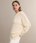 A person with straight, light brown hair wears the cream-colored Cruise Cocoon Crewneck sweater—lightweight with an oversized silhouette—and white pants, posing with one hand in their pocket against a plain beige background.