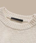 A close-up of the cream-colored Cruise Cocoon Crewneck, a lightweight knit sweater with a ribbed crewneck and oversized fit, featuring a black label sewn inside the collar against a beige background.
