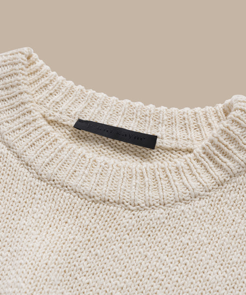 A close-up of the cream-colored Cruise Cocoon Crewneck, a lightweight knit sweater with a ribbed crewneck and oversized fit, featuring a black label sewn inside the collar against a beige background.