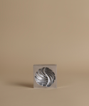 A silver metallic sculptural object with a twisted, ribbed form displayed inside a clear square acrylic block, set against a solid warm beige background.