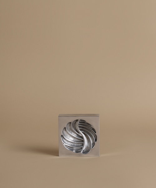 A silver metallic sculptural object with a twisted, ribbed form displayed inside a clear square acrylic block, set against a solid warm beige background.