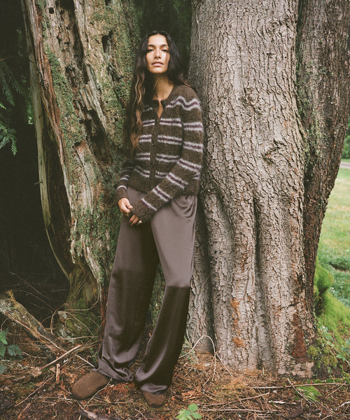A woman stands outdoors leaning against a large tree trunk, wearing a brown and gray striped knit cardigan, taupe satin pants, and brown suede boots.