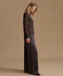 A woman stands in profile wearing a dark brown long-sleeve knit top and dark charcoal wide-leg pants against a warm beige studio background.