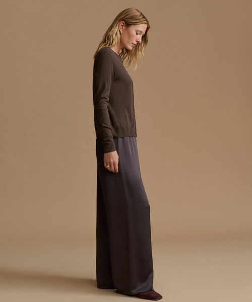 A woman stands in profile wearing a dark brown long-sleeve knit top and dark charcoal wide-leg pants against a warm beige studio background.