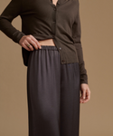 A model wearing a dark brown button-front cardigan and dark charcoal gray wide-leg trousers, shown from the chest down against a solid warm tan background.
