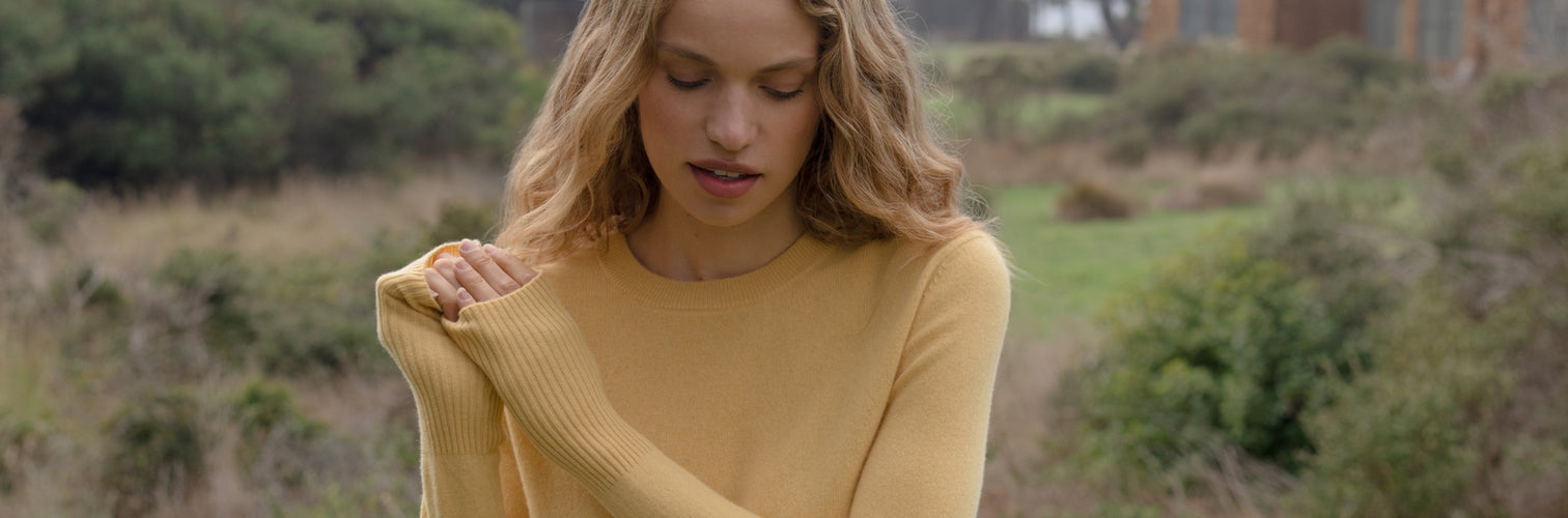 Woman in a yellow sweater standing outdoors with greenery in the background