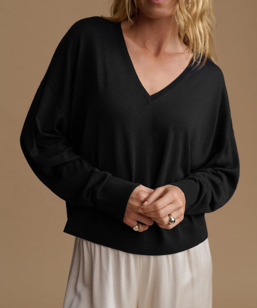 A woman wearing a black long-sleeve V-neck knit sweater with a relaxed fit, paired with light cream pants, set against a solid warm beige background.