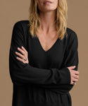 A woman wearing a black long-sleeve V-neck knit top, shown from the lips down with her arms crossed. She is wearing silver rings on her fingers.