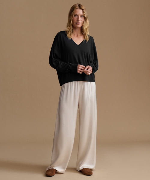 A woman modeling a black long-sleeve V-neck top and cream wide-leg trousers against a solid beige studio background.