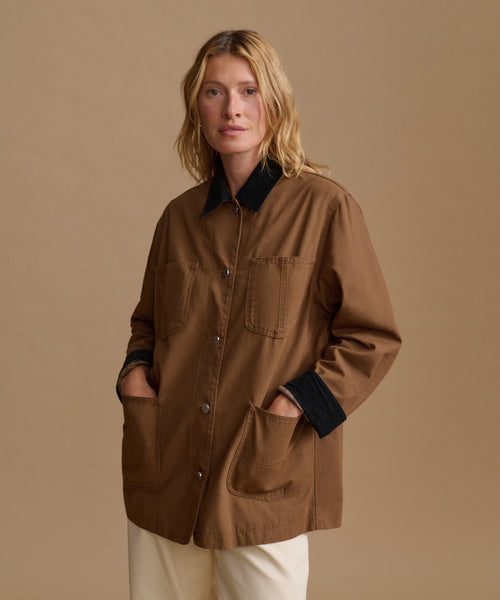 A woman modeling a brown utility-style jacket with black contrast collar, cuffs, and pocket trim, paired with cream pants, against a solid beige studio background.