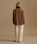 A model viewed from the back wearing a brown jacket and cream wide-leg trousers, set against a solid beige studio background.